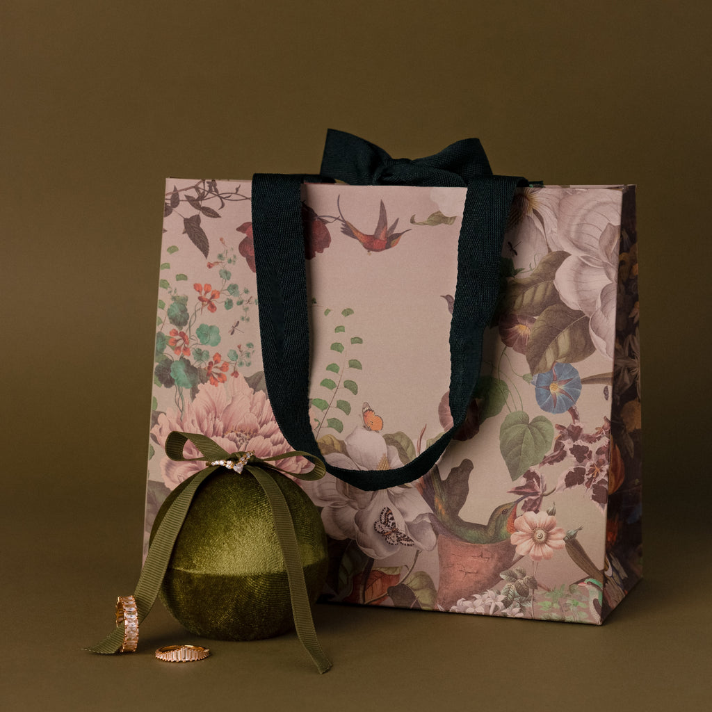 Floral gift bag with black handles, a green ornament, and two gold rings beside the Velvet Round Ring Box on an olive background.