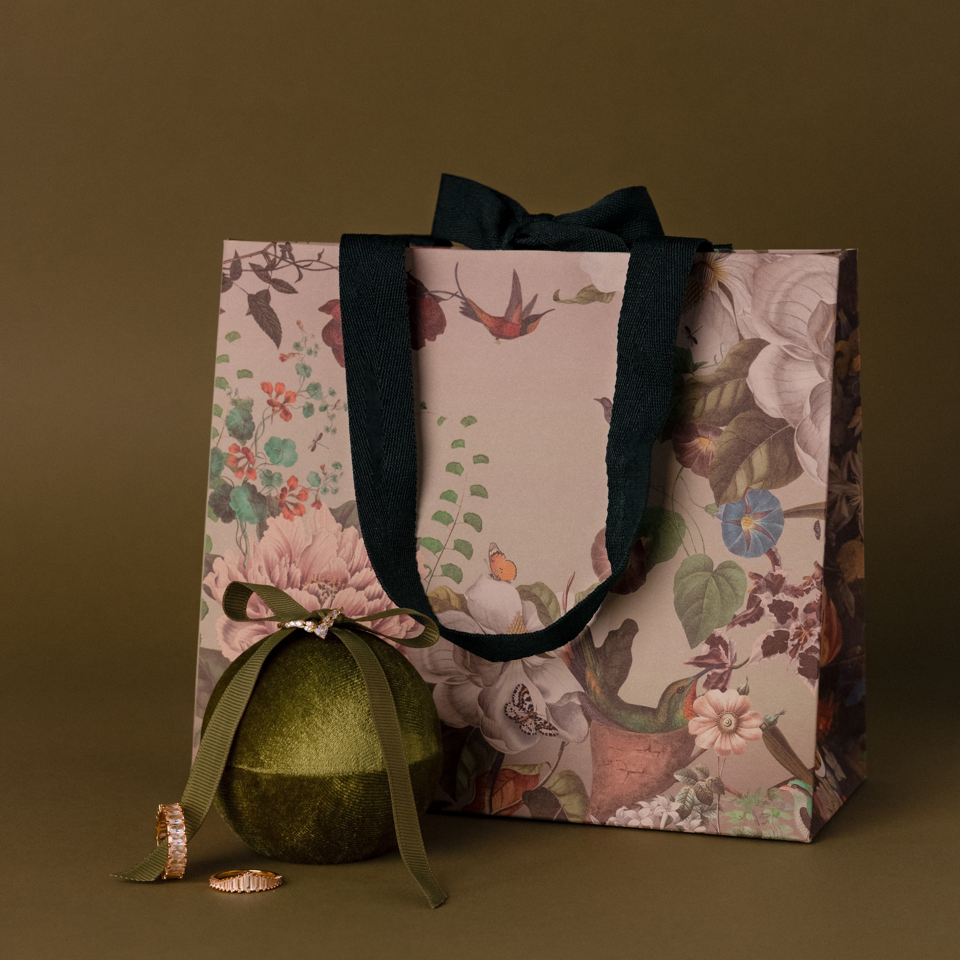 Floral gift bag with black handles, a green ornament, and two gold rings beside the Velvet Round Ring Box on an olive background.