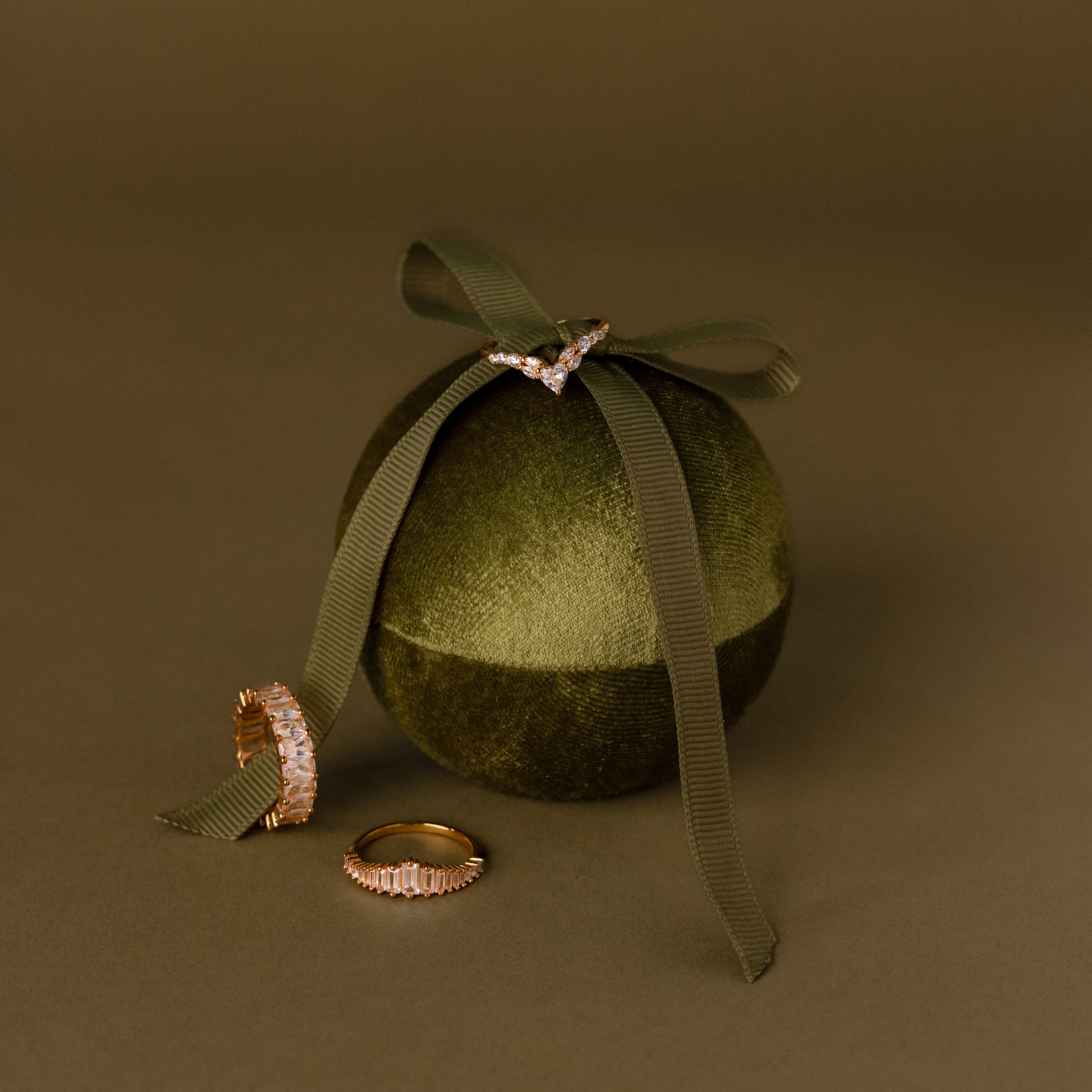 Three gold rings are displayed on and next to the Green Velvet Round Ring Box, which is elegantly tied with a matching ribbon.