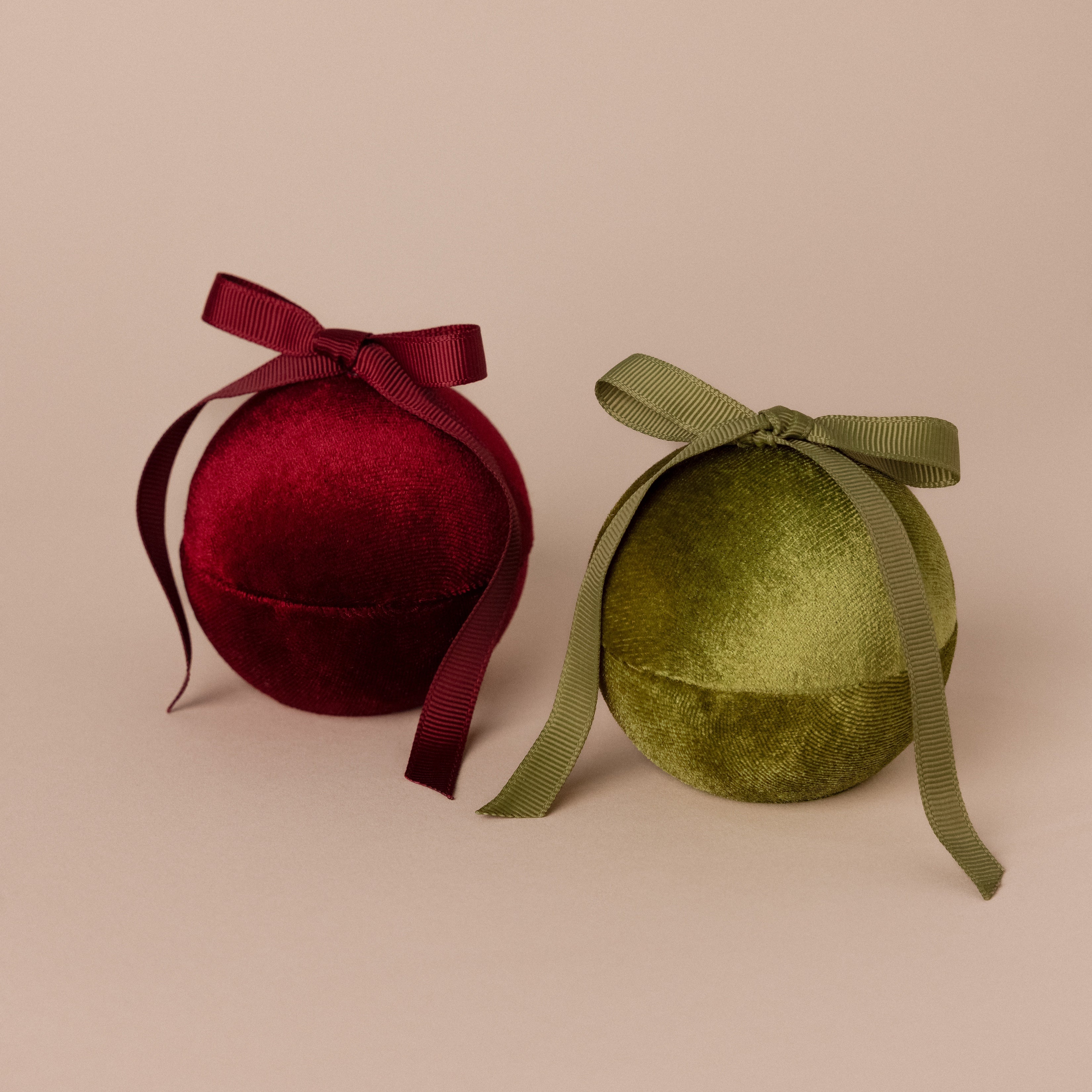 Two Velvet Round Ring Boxes in red and green, each adorned with a matching ribbon bow, are displayed elegantly on a beige background.