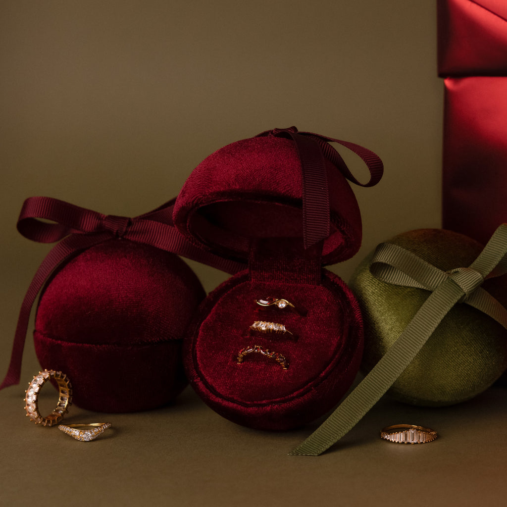 Three gold rings displayed in a Red Velvet Round Ring Box, with additional rings and ribbon-wrapped boxes nearby.