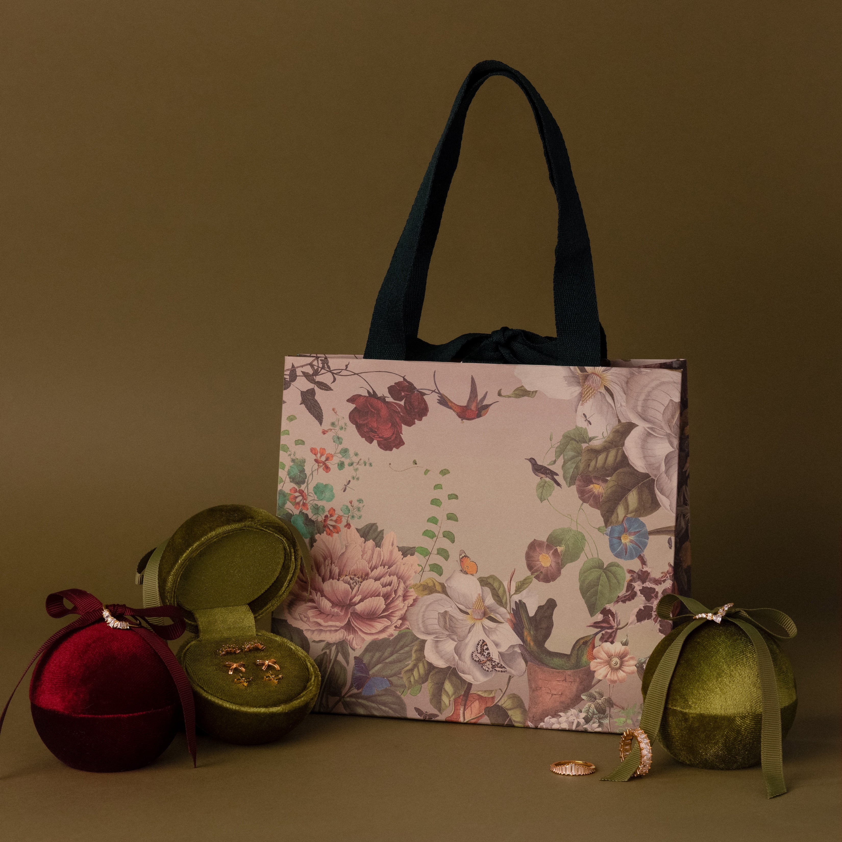 Green Velvet Round Ring Box displayed with jewelry boxes, rings, and earrings on an olive green background in a floral gift bag.