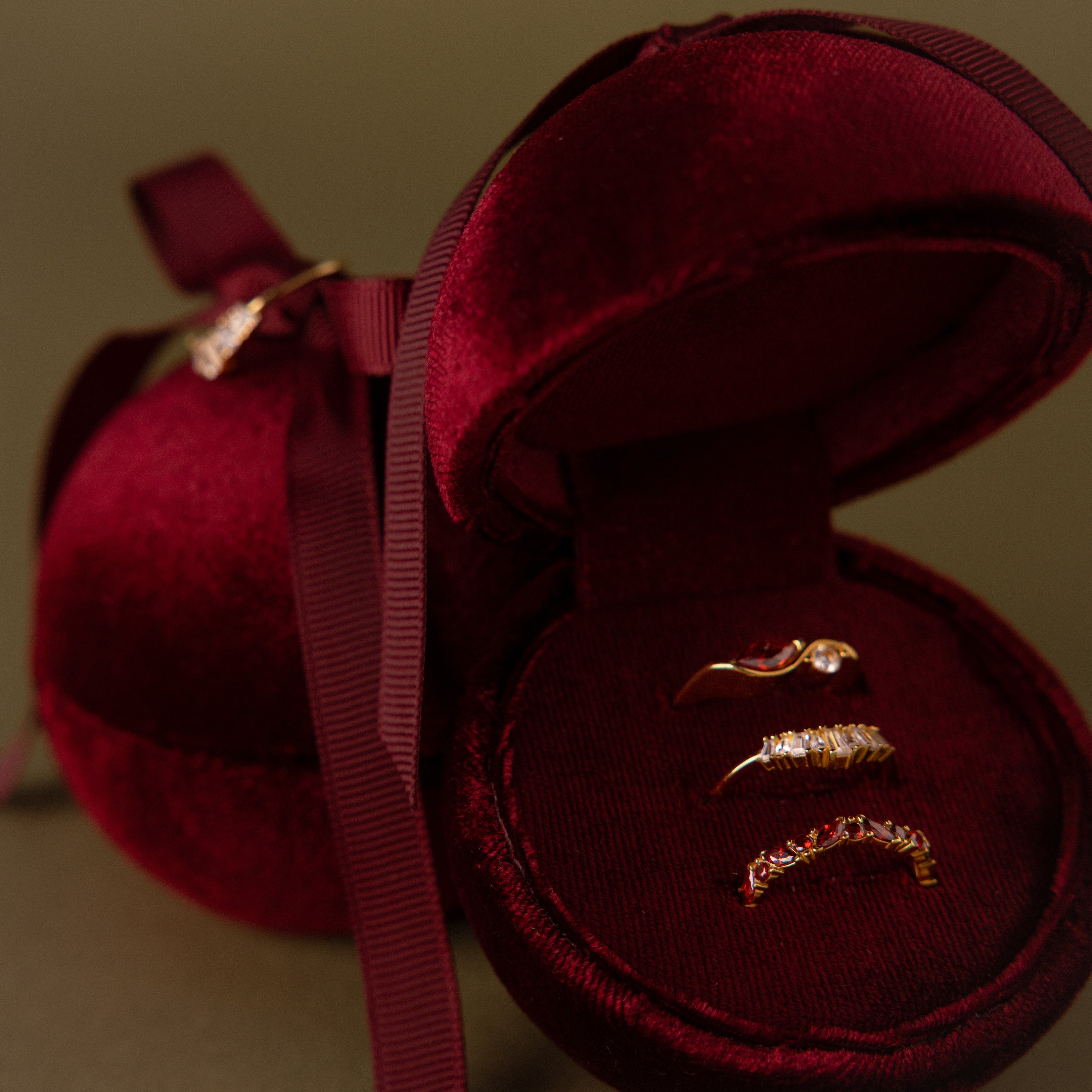 Three gold rings displayed in an open Red Velvet Round Ring Box with a matching ribbon.