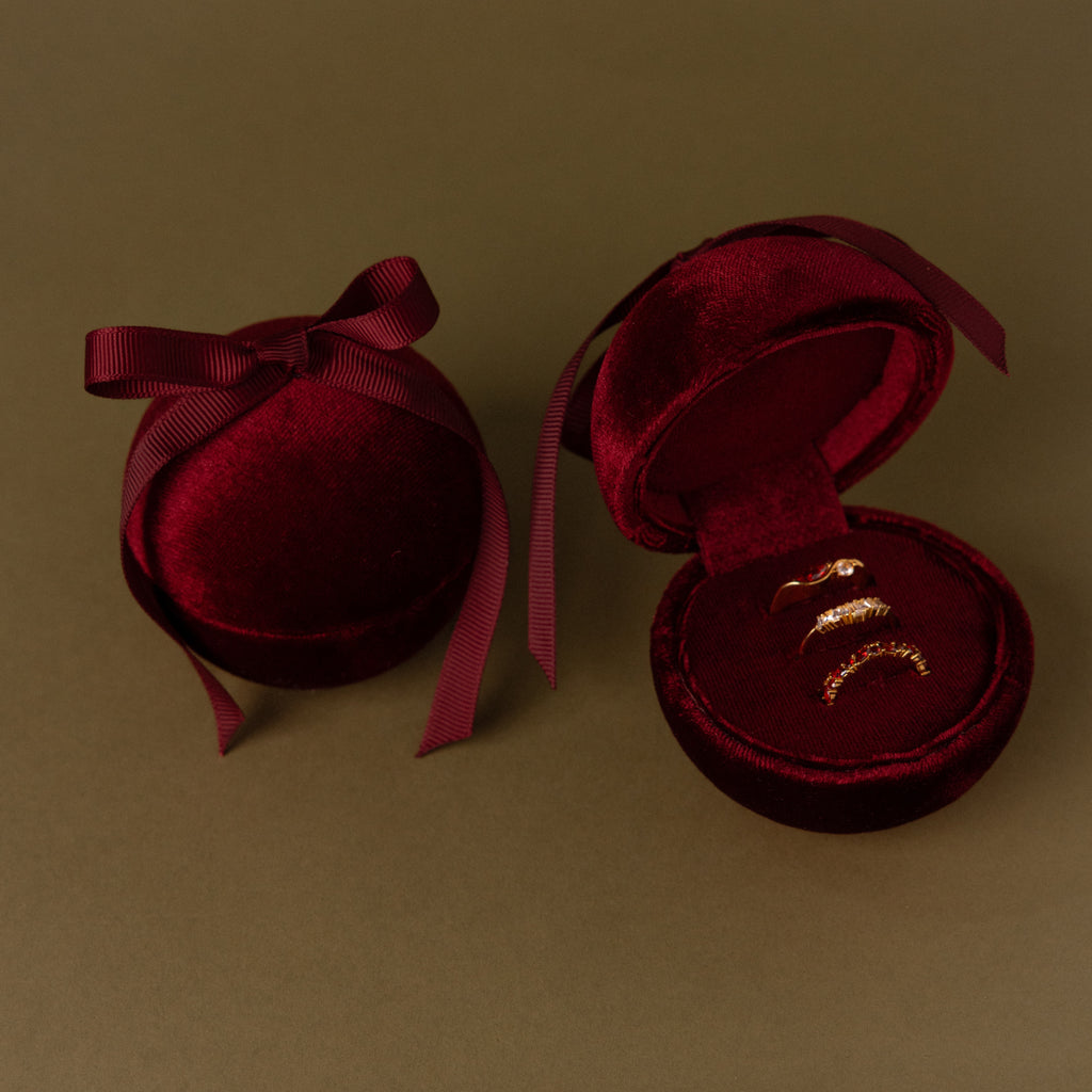 Two Red Velvet Round Ring Boxes, one open to reveal three gold rings, rest elegantly on a brown surface.