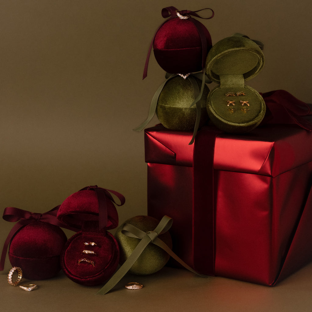 Velvet Round Ring Boxes in red and green, each holding a ring, are arranged around a large red gift box with a ribbon.