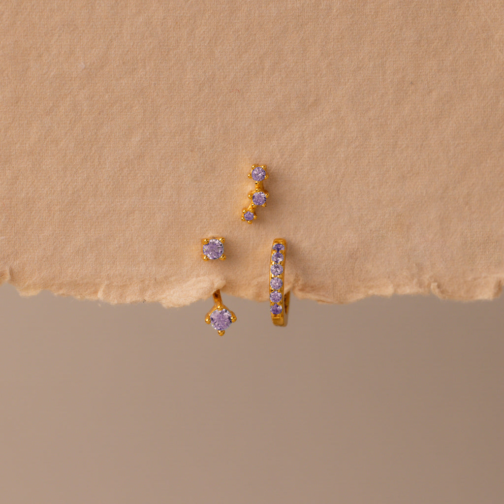 The Alexandrite Earring Trio Set features gold earrings and studs with small purple gemstones in gold settings, including a pavé huggie hoop, displayed on a textured beige surface—a chic set perfect for any occasion.