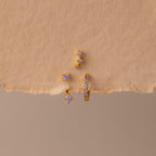 The Alexandrite Earring Trio Set features gold earrings and studs with small purple gemstones in gold settings, including a pavé huggie hoop, displayed on a textured beige surface—a chic set perfect for any occasion.