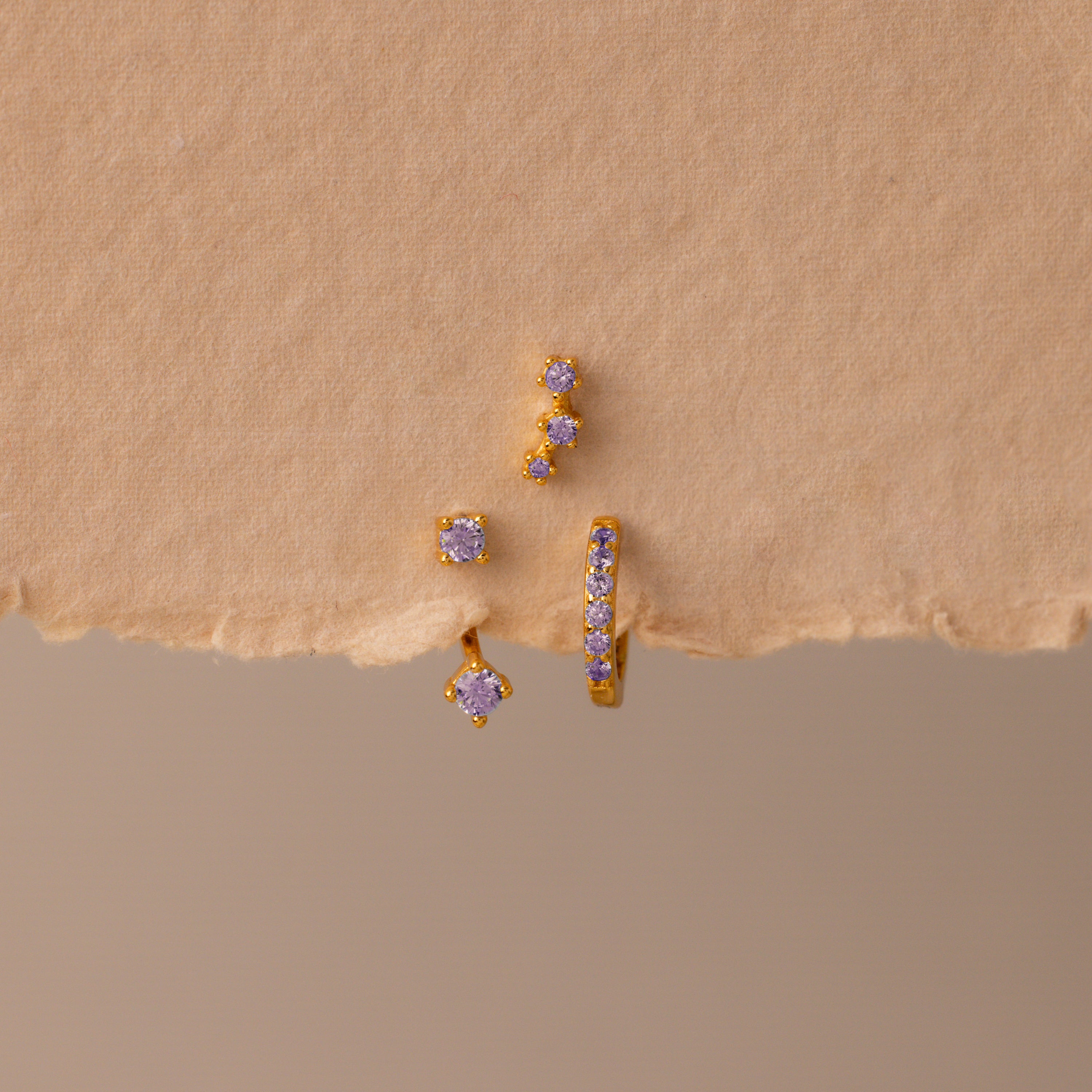 The Alexandrite Earring Trio Set features gold earrings and studs with small purple gemstones in gold settings, including a pavé huggie hoop, displayed on a textured beige surface—a chic set perfect for any occasion.