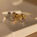Alexandrite Flatback Studs featuring gold and purple gemstones are displayed on a textured beige surface in soft light.