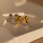 Alexandrite Flatback Studs featuring gold and purple gemstones are displayed on a textured beige surface in soft light.