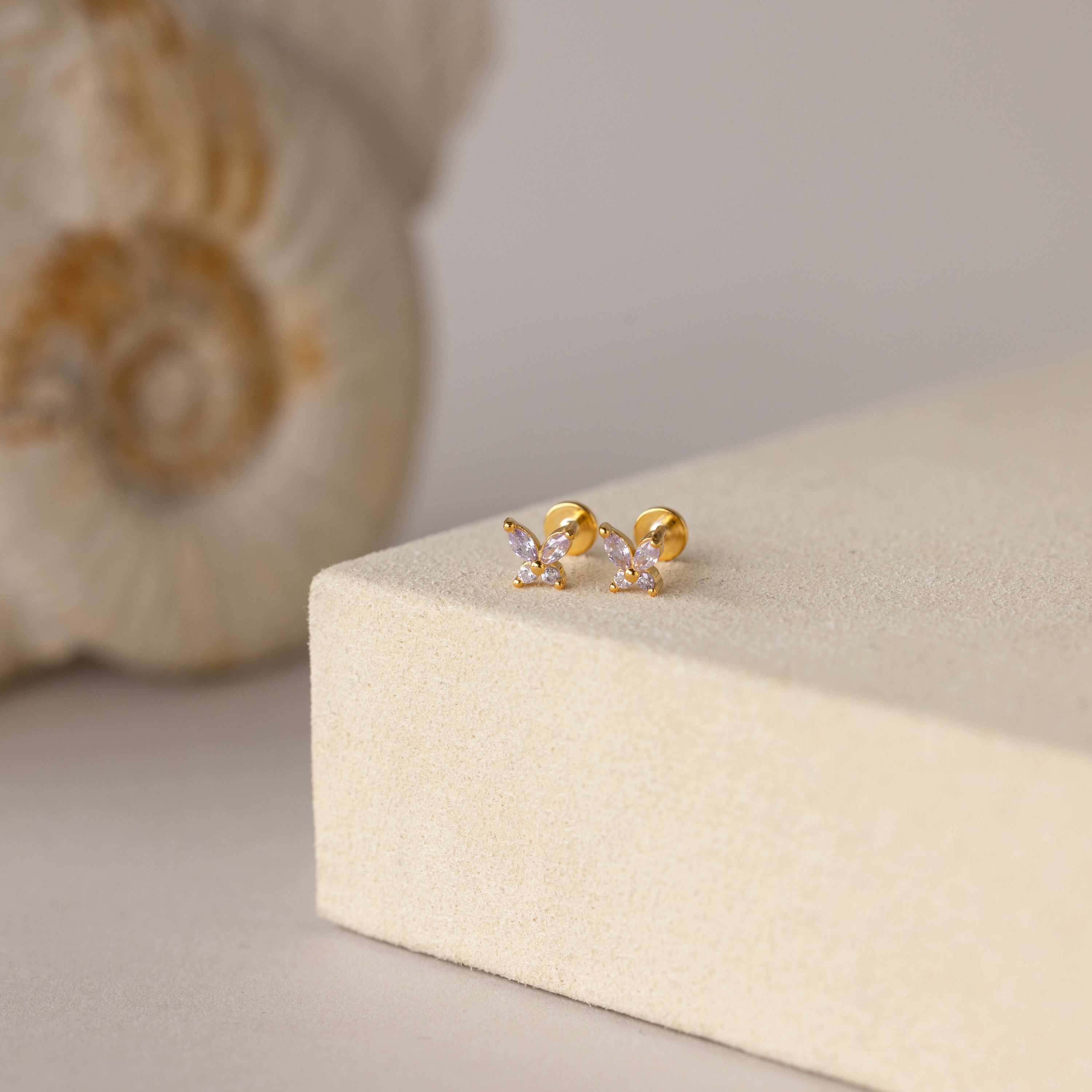 Butterfly Alexandrite Flatback Studs with small crystals, ideal as personalized jewelry, are displayed on a beige surface with a seashell in the background.