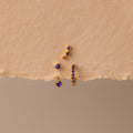 The Amethyst Earring Trio Set, featuring gold pieces with pavé huggie hoops adorned with purple stones, is displayed on a textured beige background.