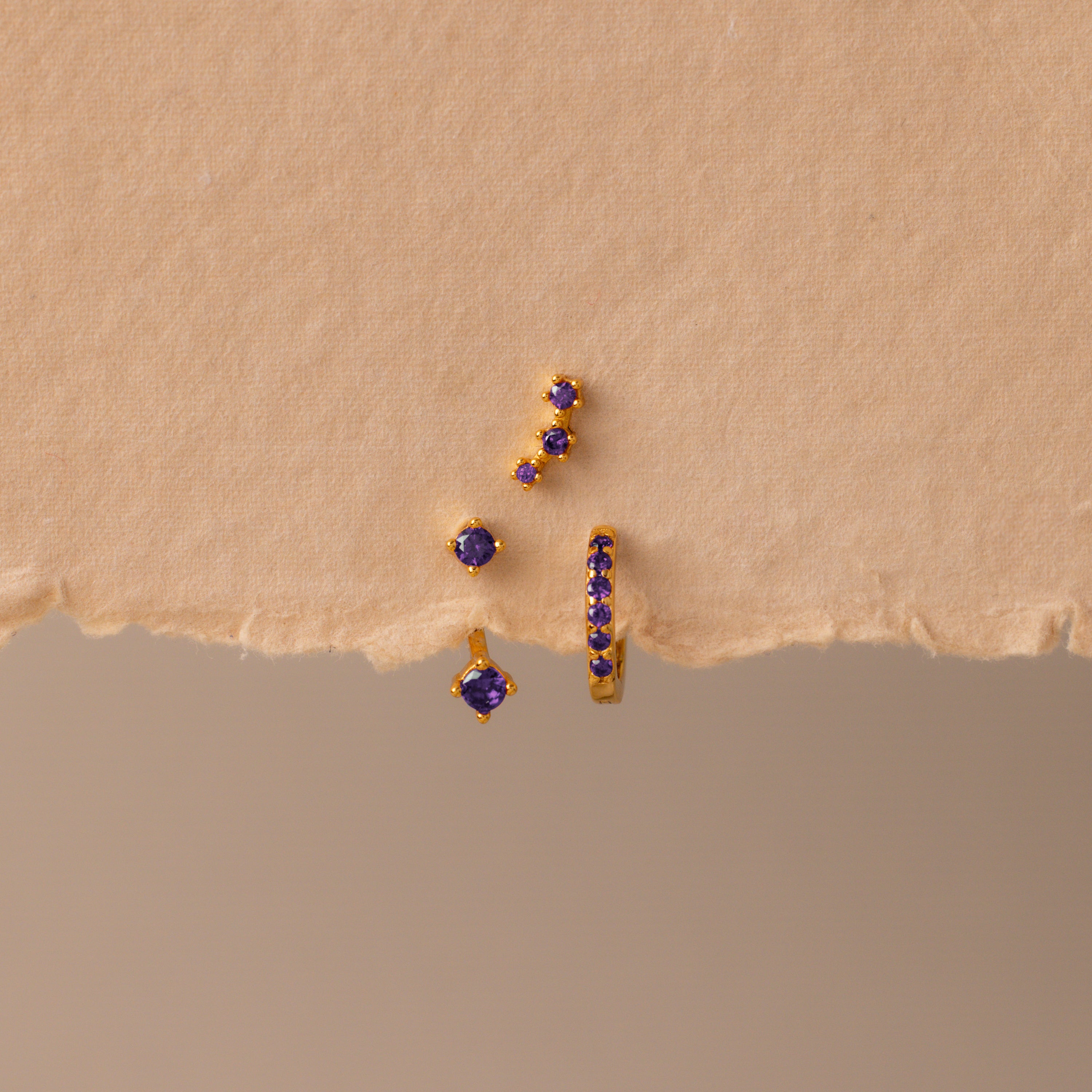 The Amethyst Earring Trio Set, featuring gold pieces with pavé huggie hoops adorned with purple stones, is displayed on a textured beige background.