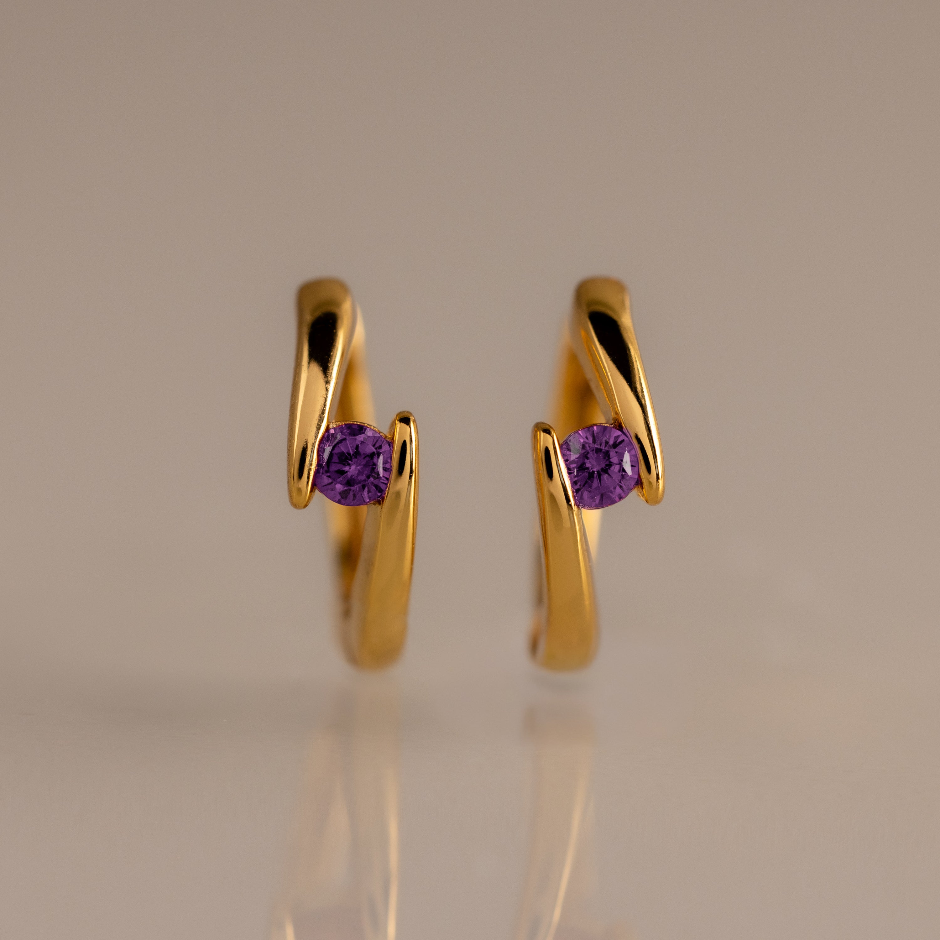 The Floating Amethyst Huggies, featuring gold earrings with a central purple gemstone, are showcased against a plain beige background.