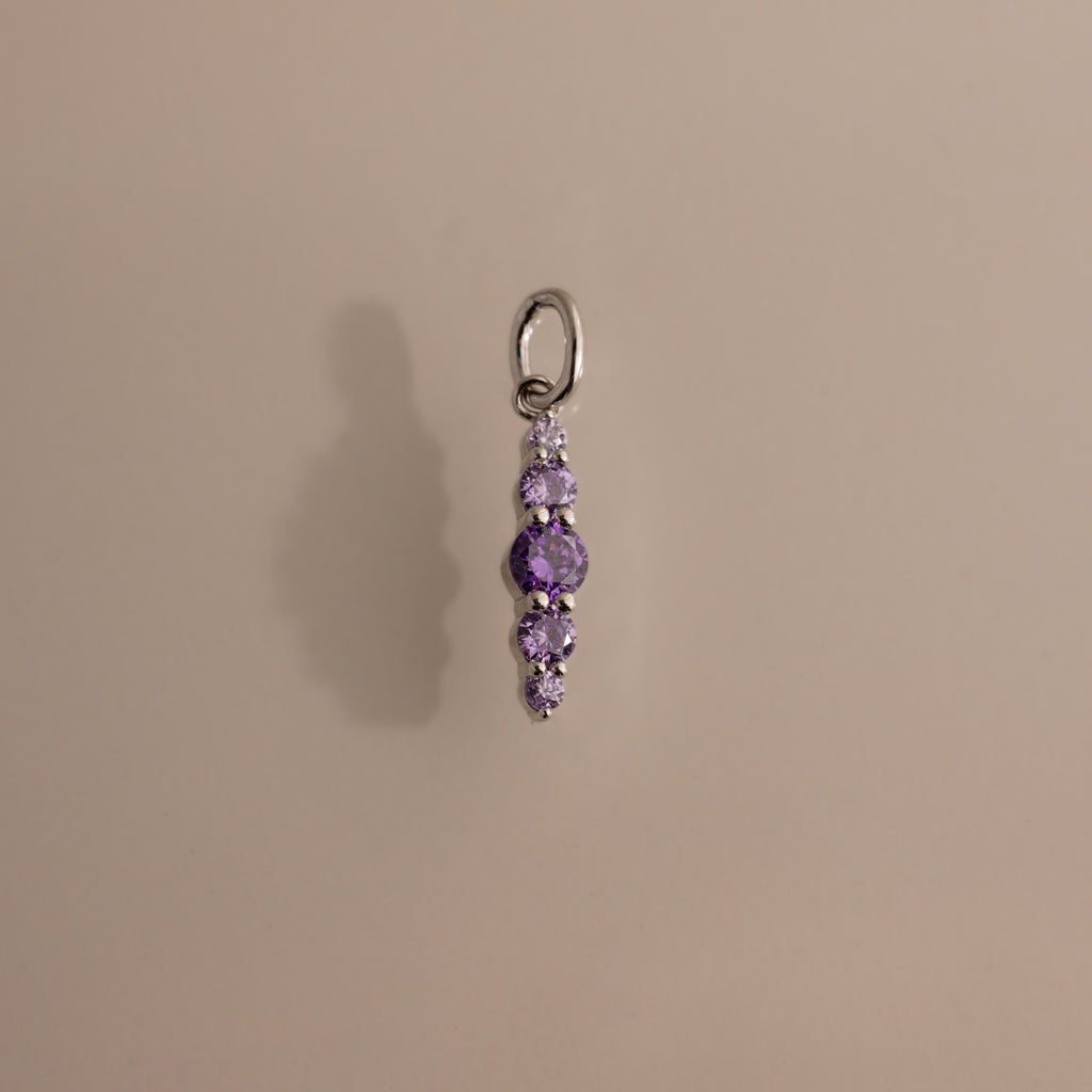 The Ombre Lace Alexandrite Charm is a silver pendant featuring five small, round purple stones arranged vertically, shown on a beige background.