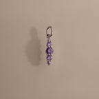 The Ombre Lace Alexandrite Charm is a silver pendant featuring five small, round purple stones arranged vertically, shown on a beige background.