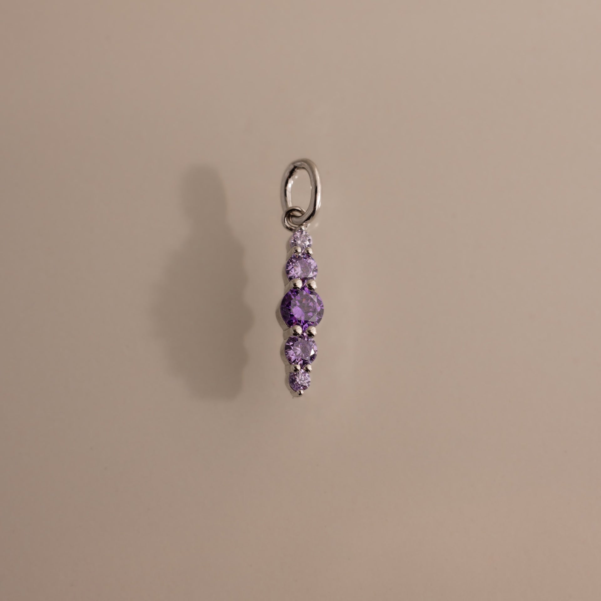 The Ombre Lace Alexandrite Charm is a silver pendant featuring five small, round purple stones arranged vertically, shown on a beige background.