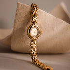 The Petite Oval Link Timepiece, a vintage-inspired gold-tone watch with a linked band, is displayed against a textured beige background.