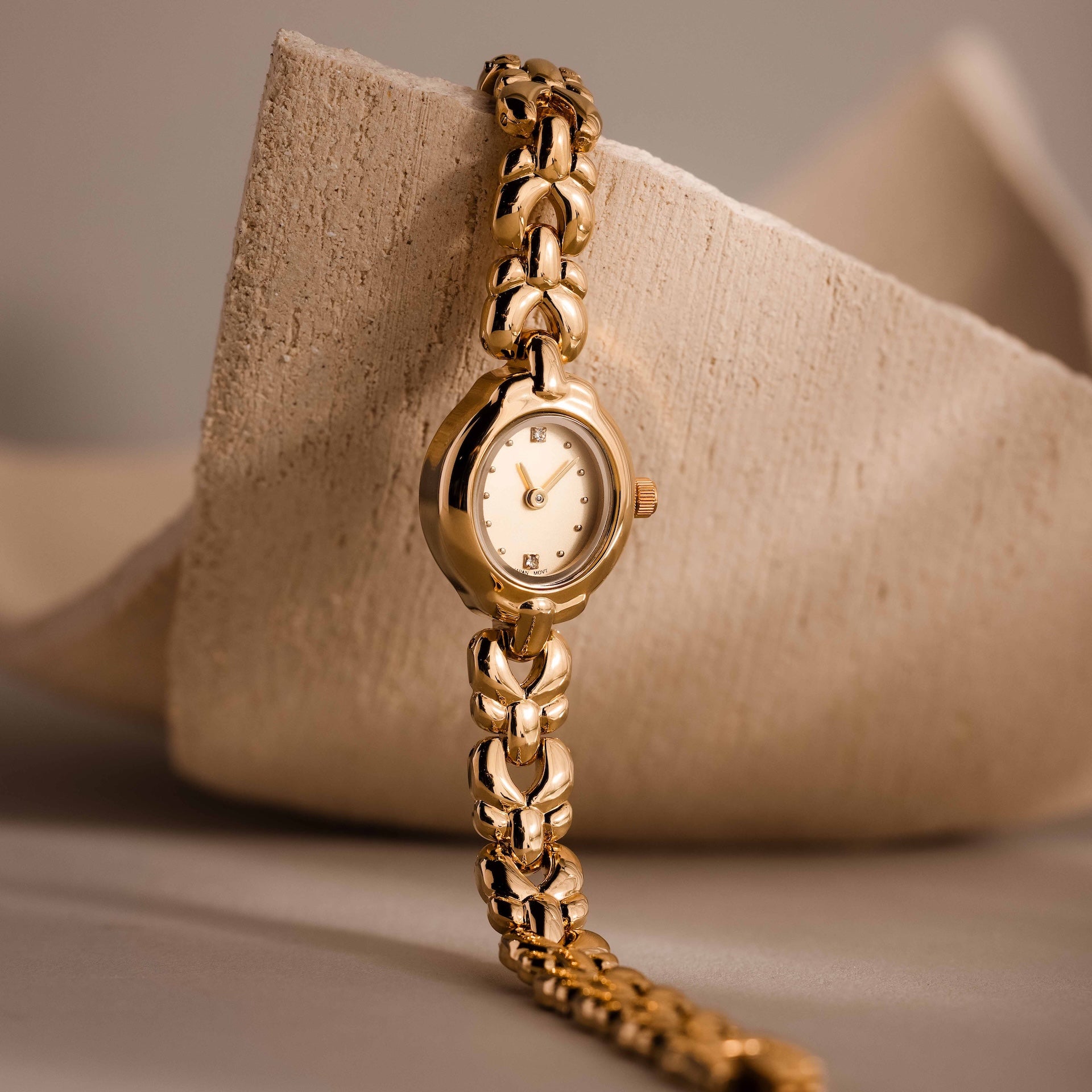 The Petite Oval Link Timepiece, a vintage-inspired gold-tone watch with a linked band, is displayed against a textured beige background.
