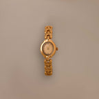 The Petite Oval Link Timepiece, a vintage-inspired gold-tone watch with a chain-link band, is displayed on a plain beige background.