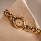 The Petite Oval Link Timepiece, a vintage-inspired gold-tone watch with a decorative chain band, rests on a textured beige stone surface.