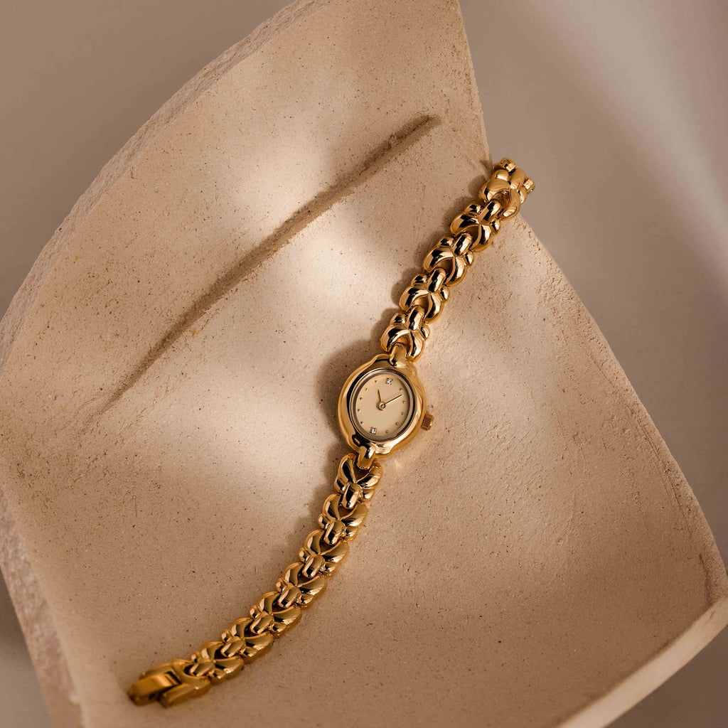 The Petite Oval Link Timepiece, a vintage-inspired gold-tone watch with a chain link band, is displayed on a textured, curved beige surface.