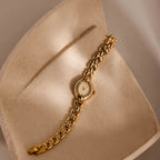 The Petite Oval Link Timepiece, a vintage-inspired gold-tone watch with a chain link band, is displayed on a textured, curved beige surface.