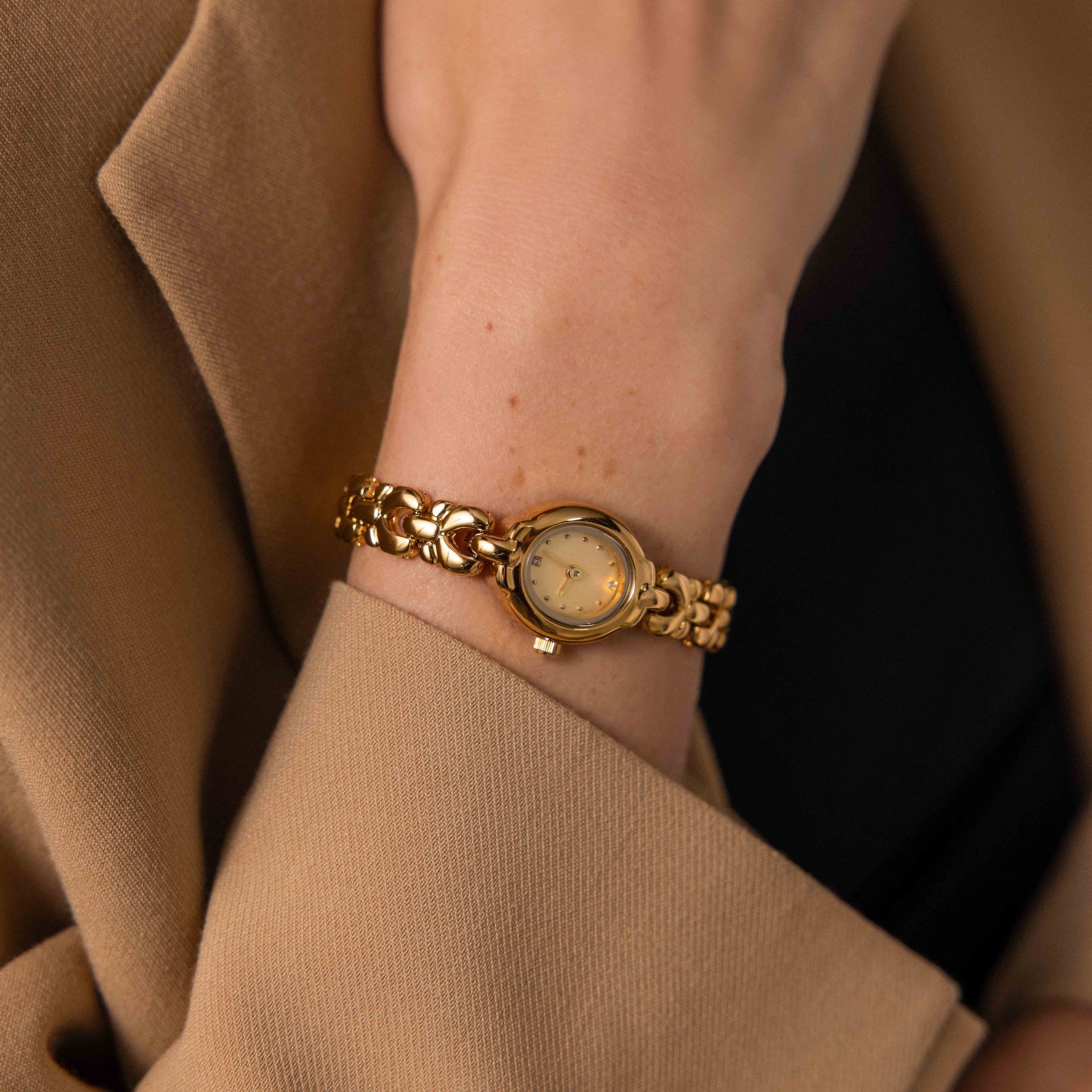 The Petite Oval Link Timepiece, a vintage-inspired gold-tone watch with a floral band, graces the wrist of someone in a beige blazer.