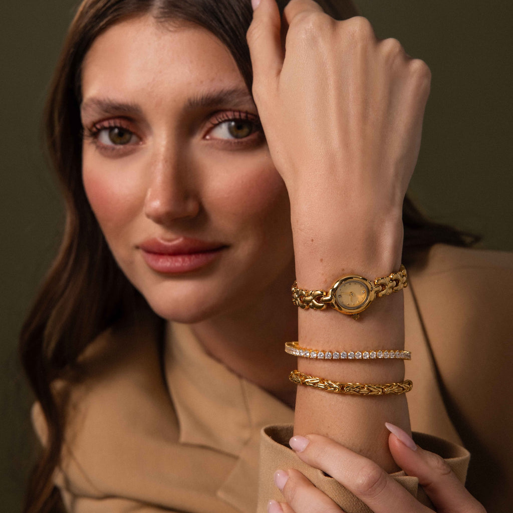 A woman in beige clothing showcases vintage-inspired gold bracelets and the Petite Oval Link Timepiece, looking at the camera.