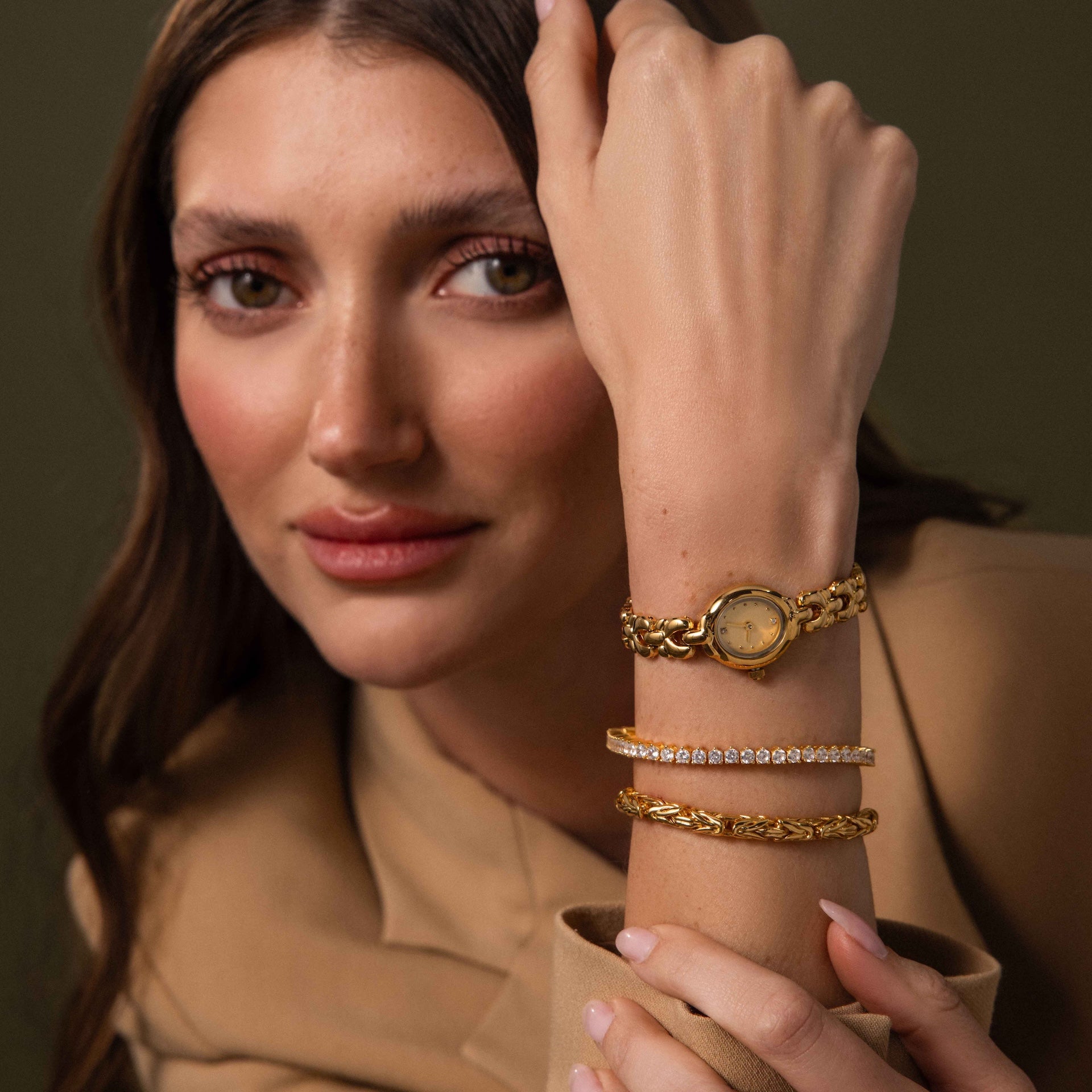 A woman in beige clothing showcases vintage-inspired gold bracelets and the Petite Oval Link Timepiece, looking at the camera.