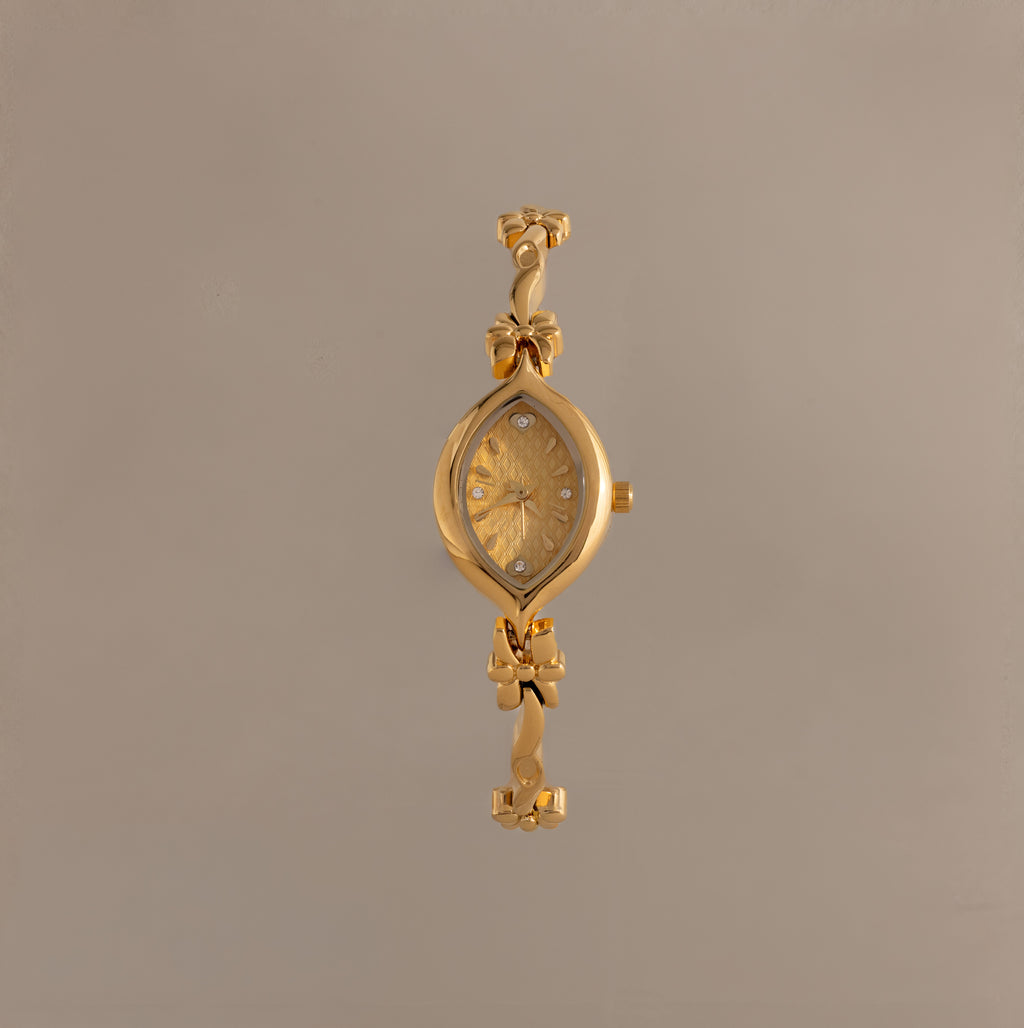 The Floral Twist Timepiece is a gold-tone bracelet watch with a floral-patterned face, marquise-shaped dial, and decorative bow accents on the band.