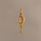 The Floral Twist Timepiece is a gold-tone bracelet watch with a floral-patterned face, marquise-shaped dial, and decorative bow accents on the band.