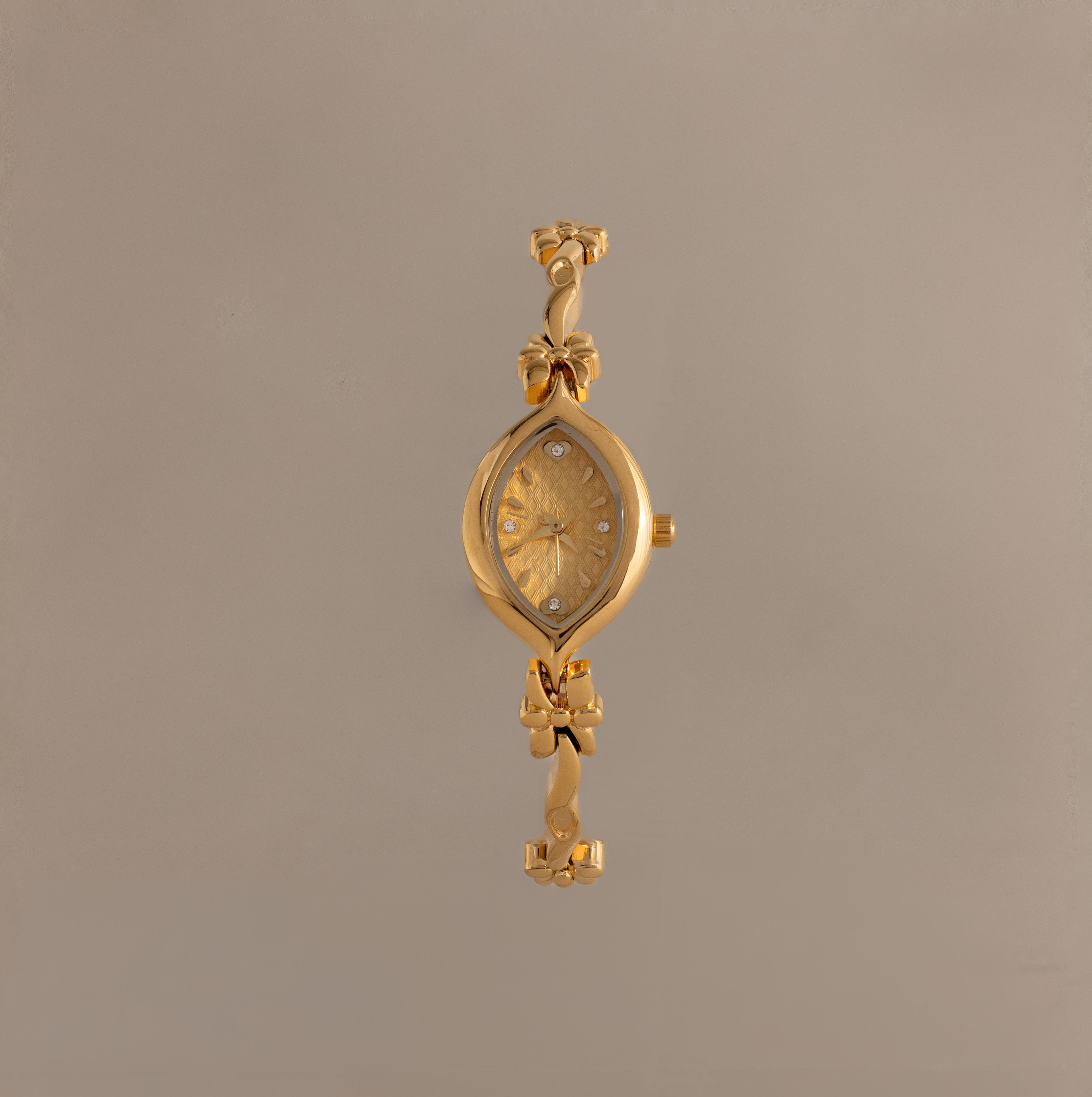 The Floral Twist Timepiece is a gold-tone bracelet watch with a floral-patterned face, marquise-shaped dial, and decorative bow accents on the band.