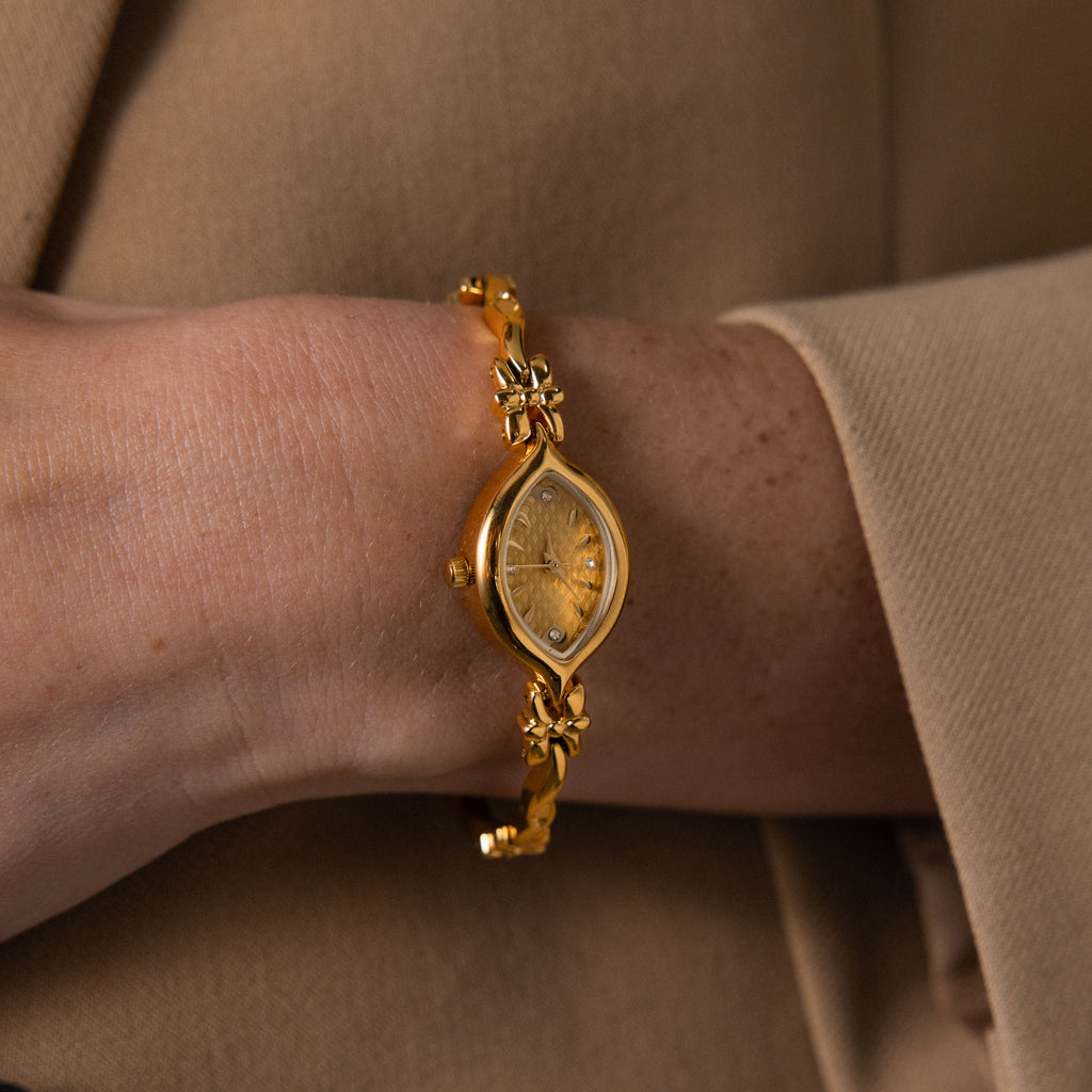 The Floral Twist Timepiece, with a gold tone, marquise dial, and floral band, is worn over a beige long-sleeve garment.