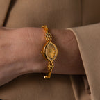 The Floral Twist Timepiece, with a gold tone, marquise dial, and floral band, is worn over a beige long-sleeve garment.
