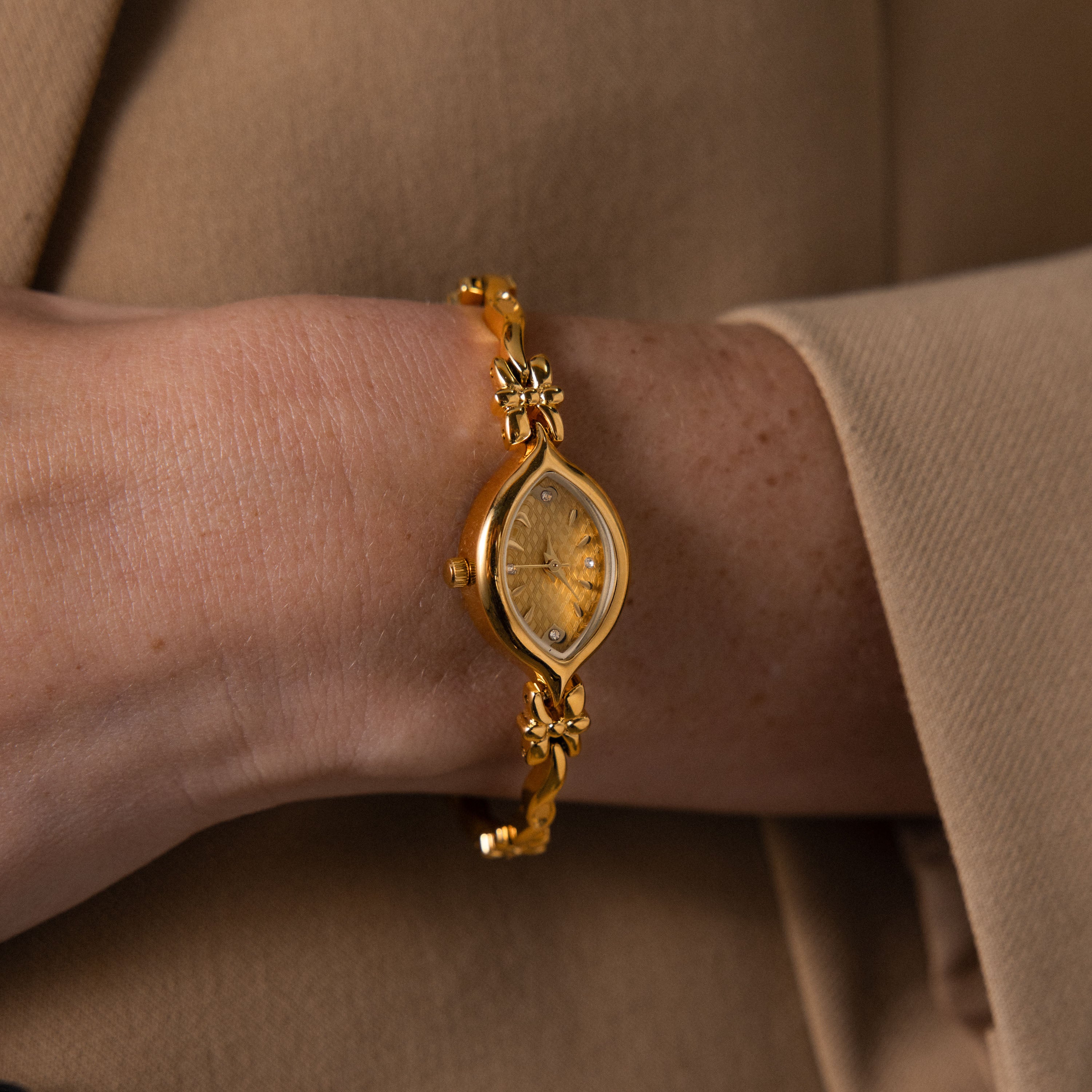 The Floral Twist Timepiece, with a gold tone, marquise dial, and floral band, is worn over a beige long-sleeve garment.