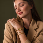 A woman in a beige blazer smiles with closed eyes, displaying gold bracelets and her elegant Floral Twist Timepiece, its marquise-shaped dial gleaming on her wrist.