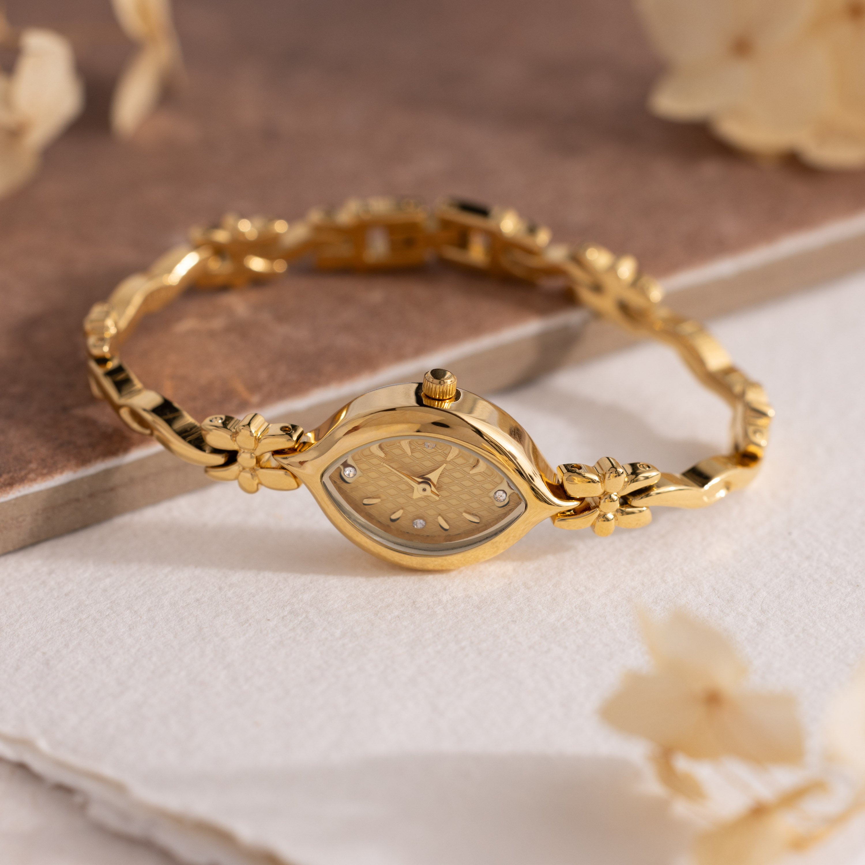 The Floral Twist Timepiece, featuring a gold tone and marquise-shaped dial, is displayed on a soft white surface amid delicate dried flowers.