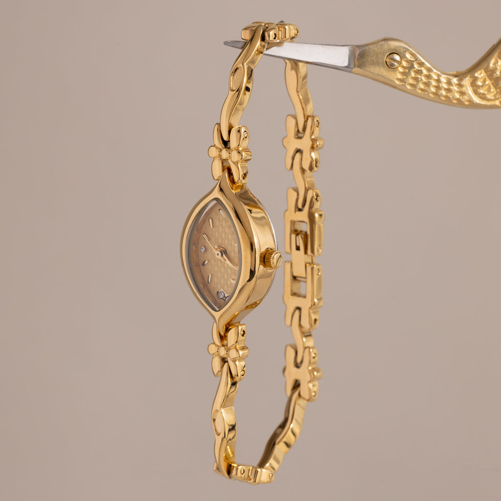 The Floral Twist Timepiece features a marquise dial with a floral design and is secured by a gold bird-shaped clamp, shown against a beige background.