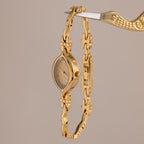 The Floral Twist Timepiece features a marquise dial with a floral design and is secured by a gold bird-shaped clamp, shown against a beige background.