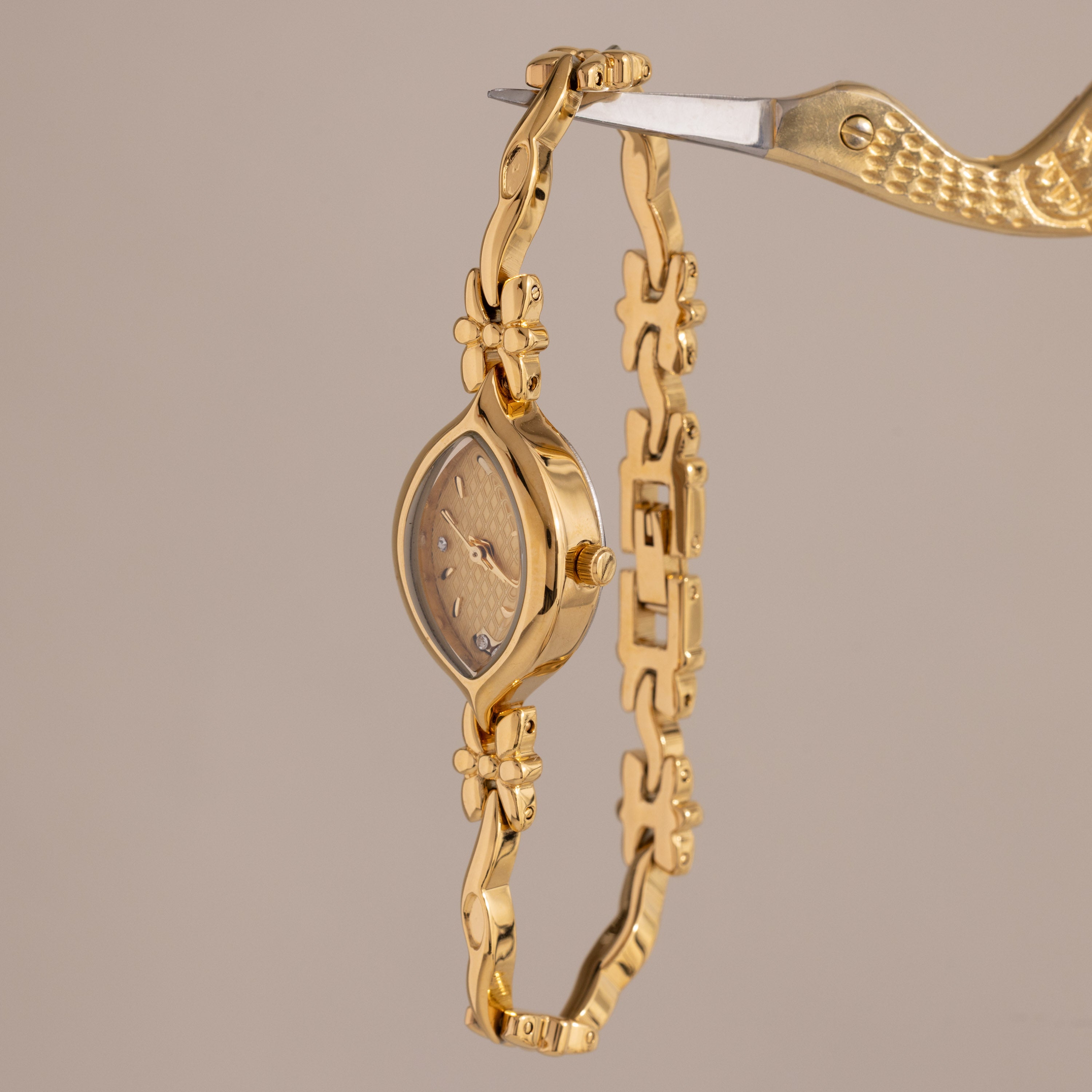 The Floral Twist Timepiece features a marquise dial with a floral design and is secured by a gold bird-shaped clamp, shown against a beige background.