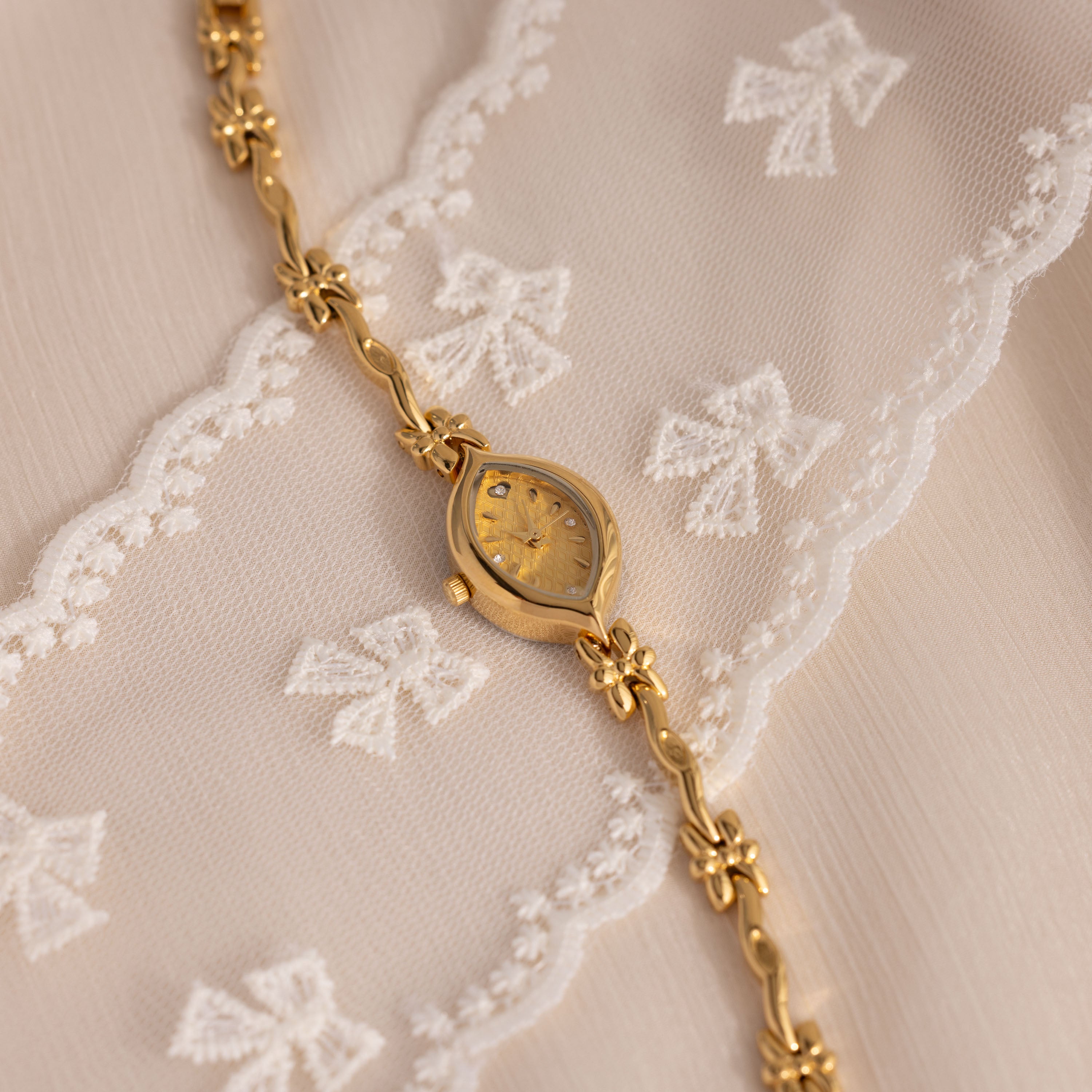 The Floral Twist Timepiece, featuring a marquise-shaped dial and gold-tone band, is elegantly displayed on white lace fabric decorated with bow patterns.
