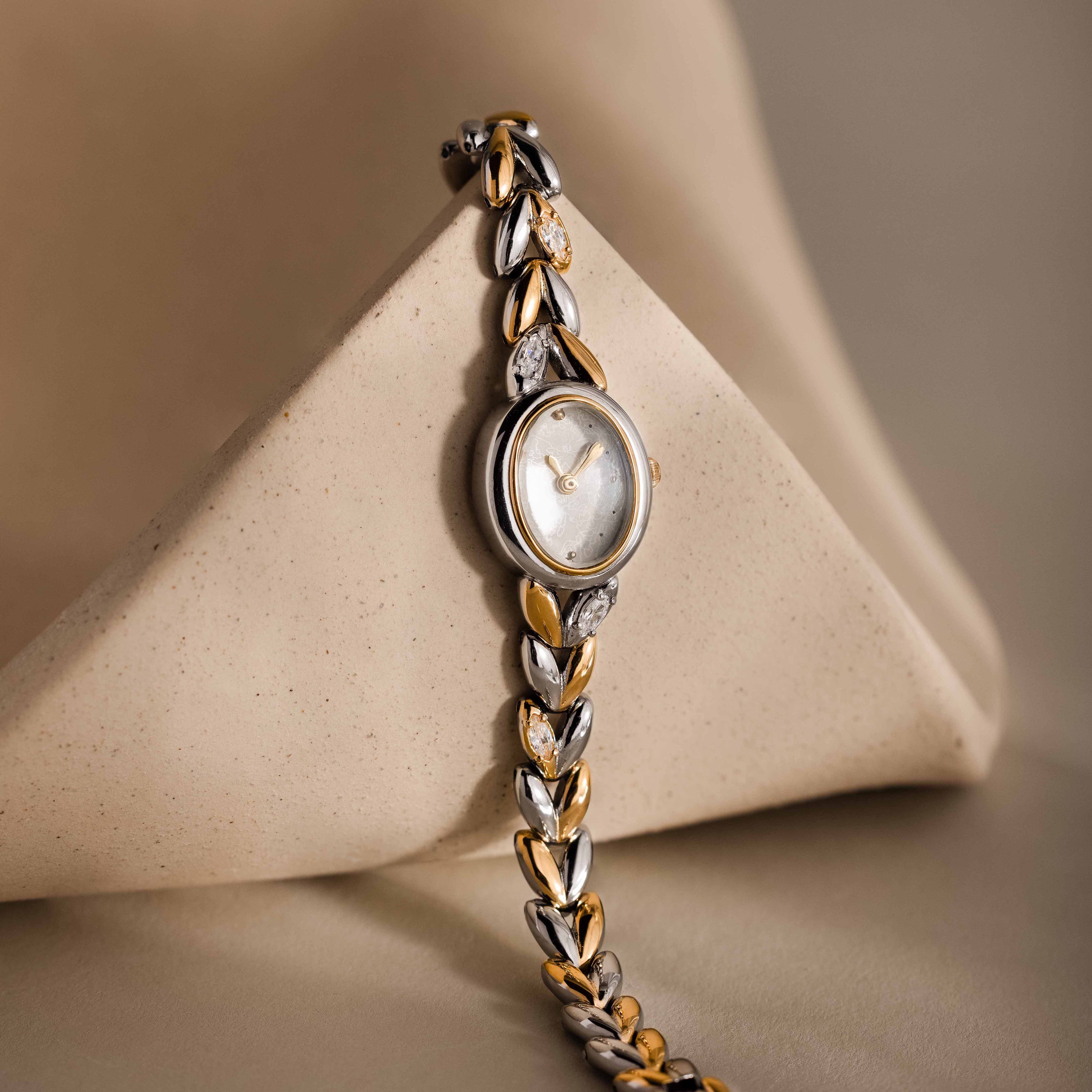 The Chevron Diamond Link Timepiece, featuring a gold-tone face and silver chain-link band, rests elegantly against a beige geometric shape.