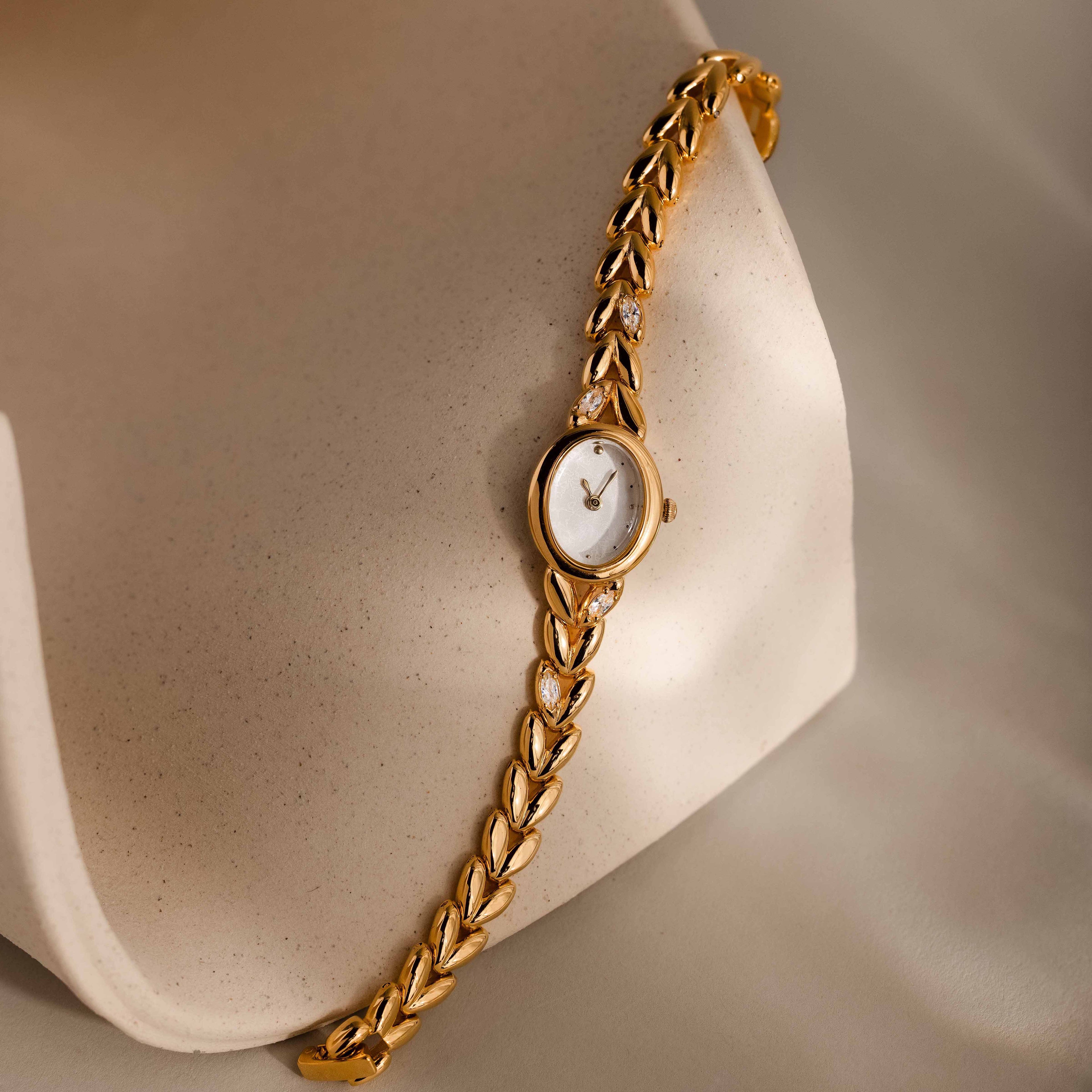 Chevron Diamond Link Timepiece in 18K Gold with a leaf-patterned band, elegantly displayed on a smooth, curved cream-colored surface.