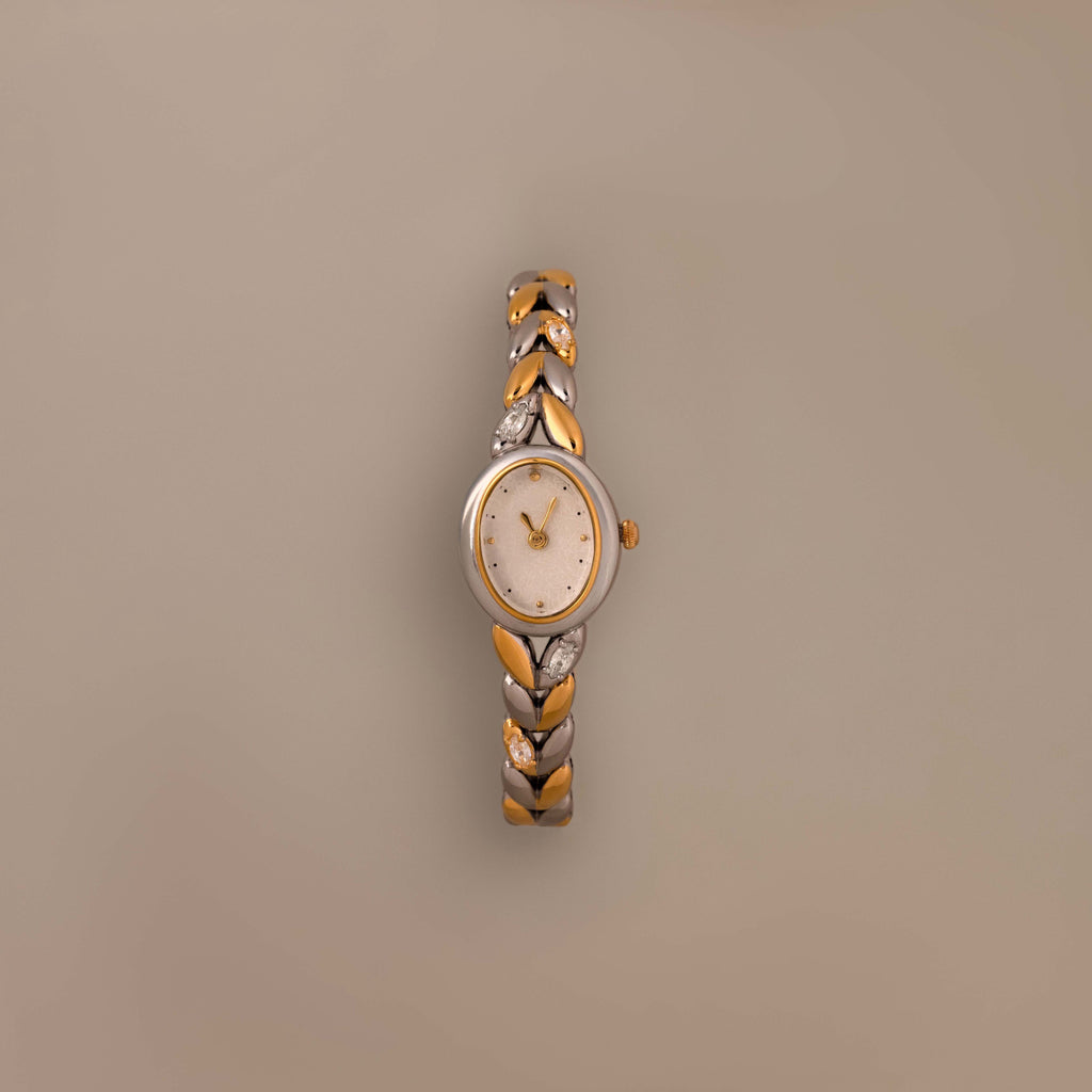 The Chevron Diamond Link Timepiece in Mixed Metal, featuring a round face and chevron-patterned band, is displayed on a plain beige background.