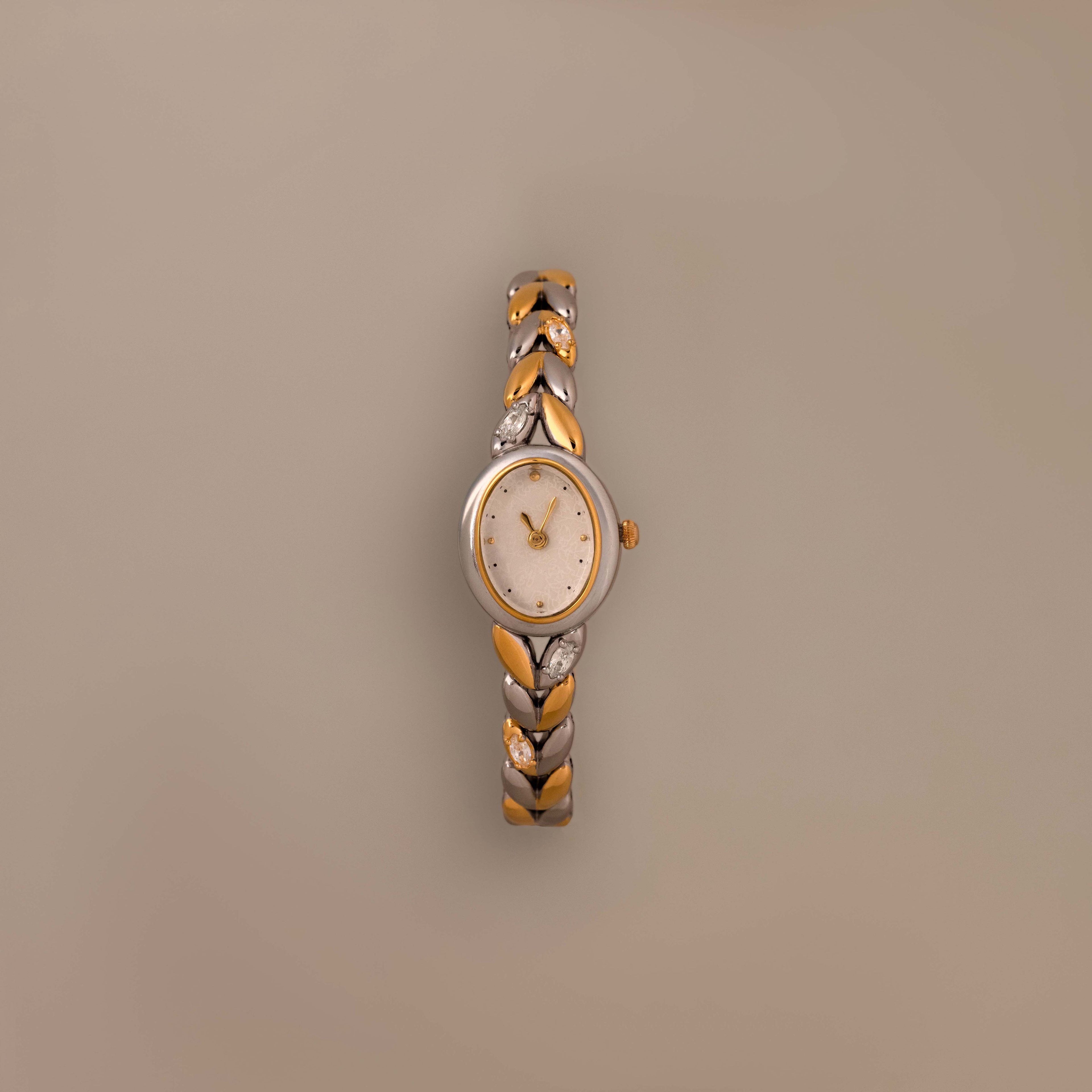 The Chevron Diamond Link Timepiece in Mixed Metal, featuring a round face and chevron-patterned band, is displayed on a plain beige background.
