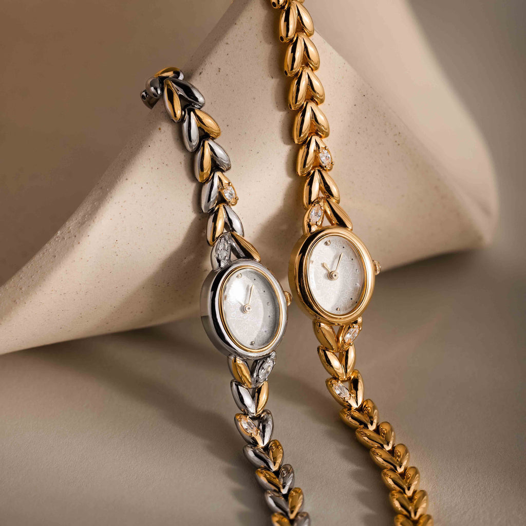 The Chevron Diamond Link Timepiece, an elegant wristwatch with a silver chevron design and leaf-pattern band, is beautifully displayed on a beige surface.