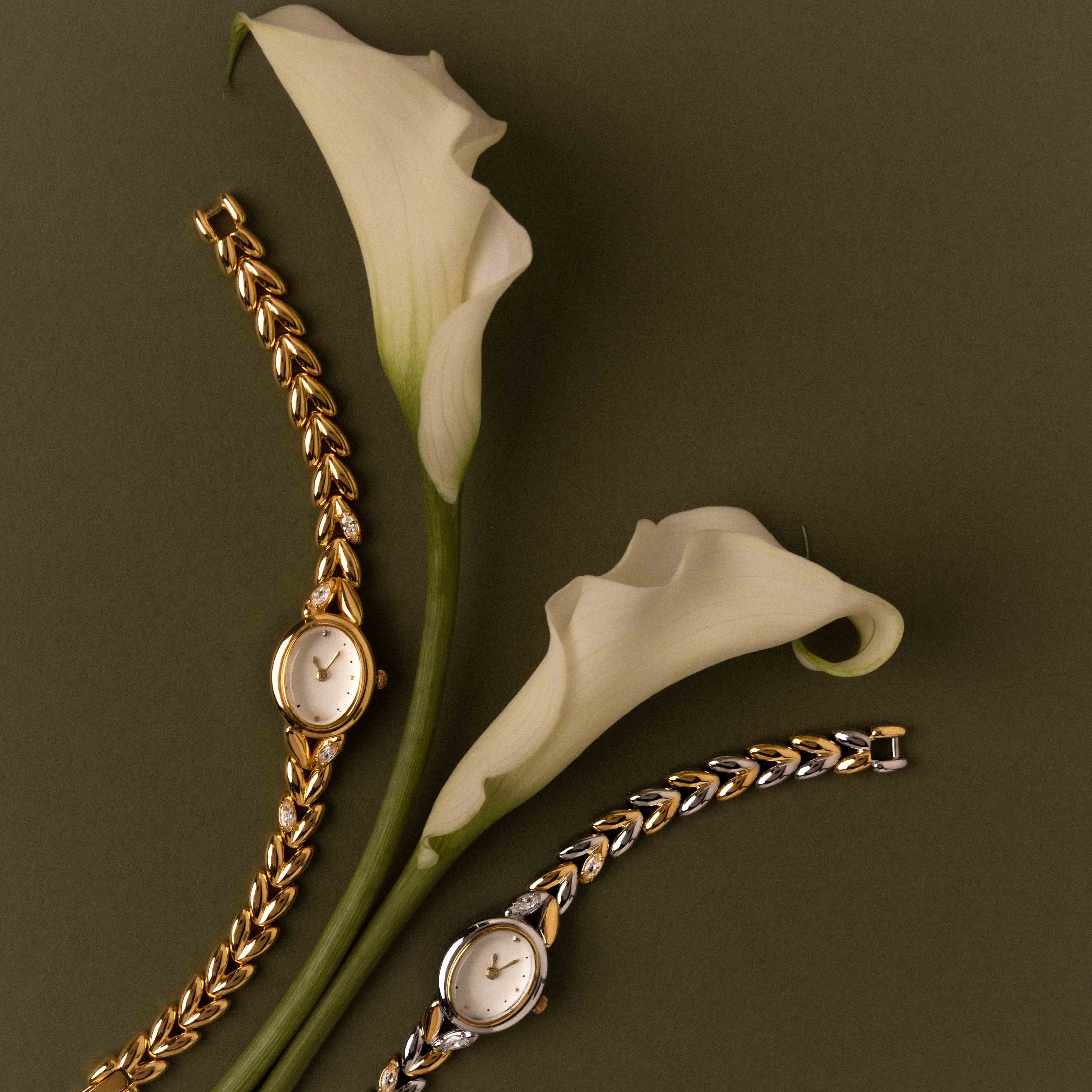 The Chevron Diamond Link Timepiece is displayed with a gold-tone watch and two white calla lilies on an olive green background.