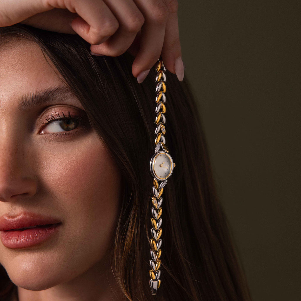 A woman holds the Chevron Diamond Link Timepiece in Mixed Metal, a stunning watch with a diamond-accented bracelet, near her face against a dark background.