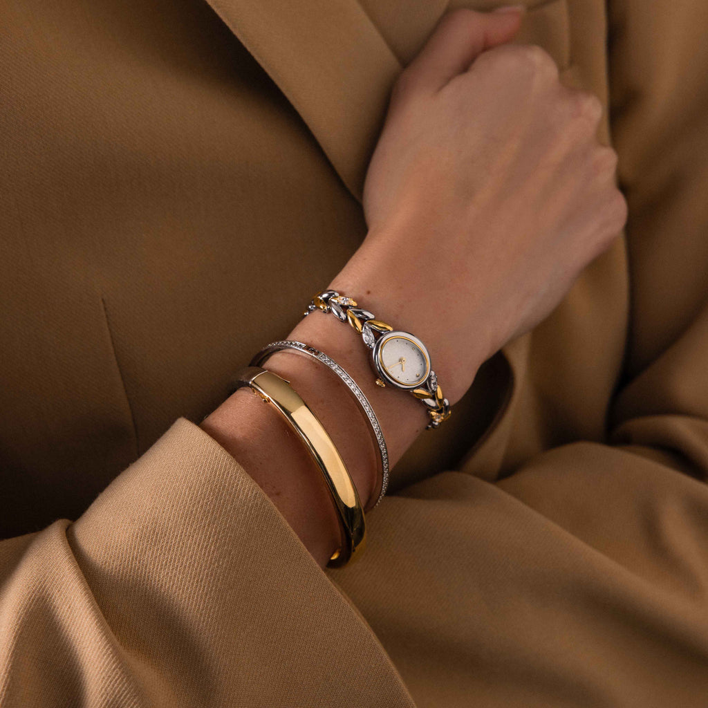 Wearing a tan coat, this person showcases the Chevron Diamond Link Timepiece—a gold-tone watch and two gold bracelets that enhance their sophisticated style.